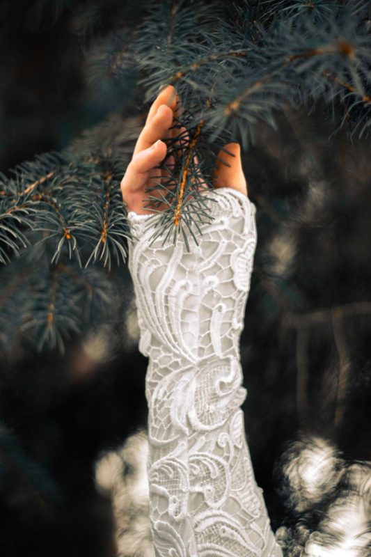 A hand in front of a pine tree, detailing the lace of a wedding dress sleeve